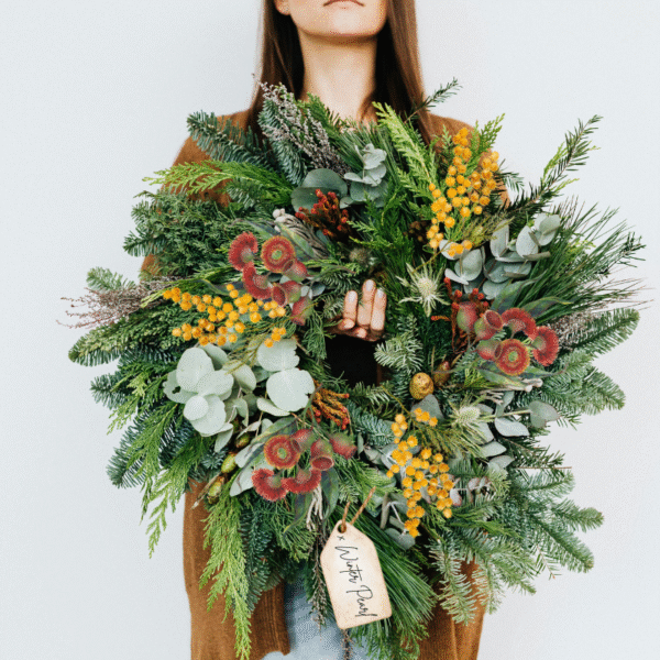 A woman with light skin and brown hair is holding a large beautiful wreath in front of her. The wreath is made from Australian native plants including yellow banksia flowers, red gumnuts and green eucalyptus leaves. It has a tag hanging at the bottom that reads "x Winter Pearl"
