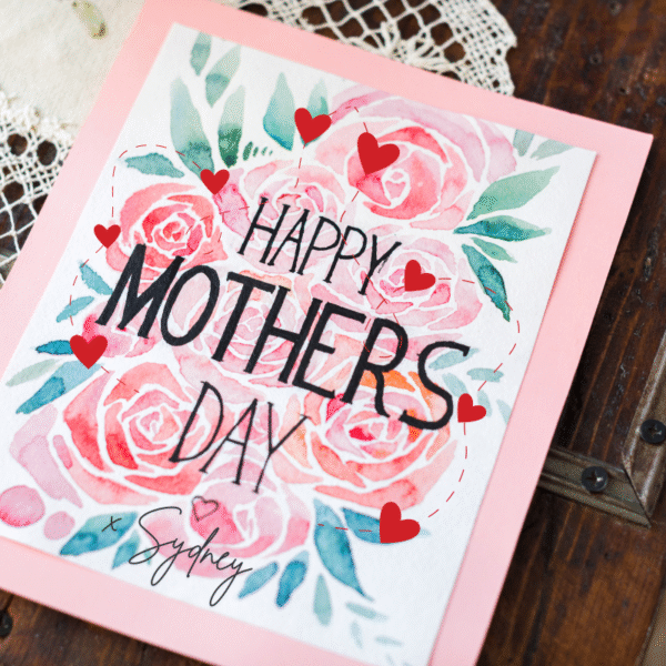 This image has a card that was created on a computer and then printed. It has a pink background and white centre with the words "Happy Mothers Day x Sydney" The words are printed on a background of pink roses, small red love hears and some green leaves. The card is sitting on a cream crocheted table runner and wooden table.